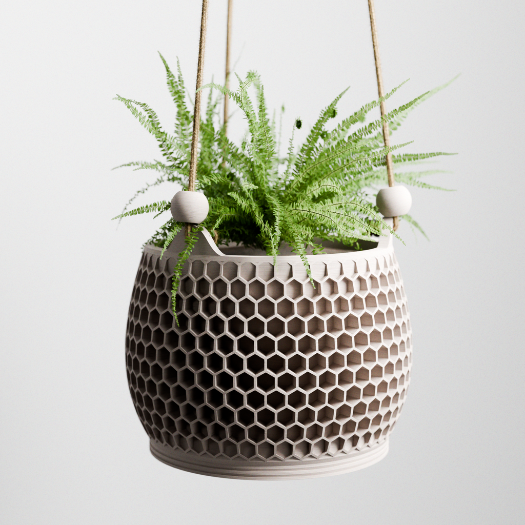 Hanging Honeycomb Planter in matte cream with geometric hexagonal cutout pattern, suspended by jute cord with ceramic bead accents, holding a lush fern against a light grey background