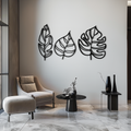 Set of three black metal laser-cut botanical wall art panels featuring a banana leaf, a philodendron leaf, and a monstera leaf, mounted on a light grey wall above a lounge chair in a modern interior